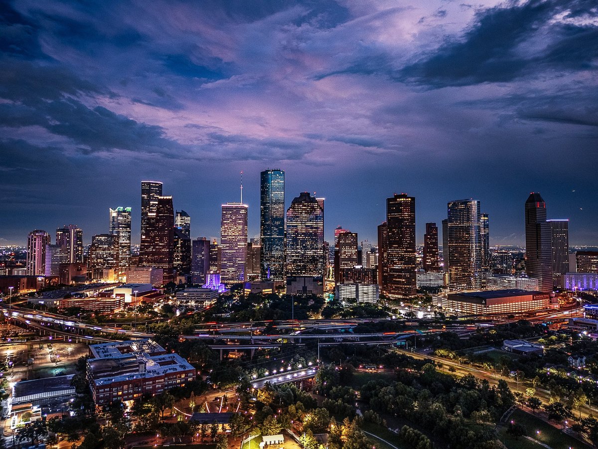 Drone aerial image of downtown Houston, Texas captured at twilight. The photograph highlights the illuminated skyscrapers, highways, and cityscape under a dramatic evening sky.